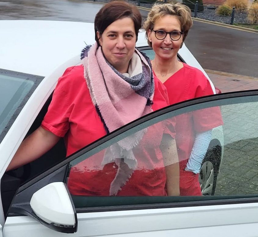 Unser Team Zwei Frauen in roten Uniformen stehen neben einem Auto, eine von ihnen lächelt.