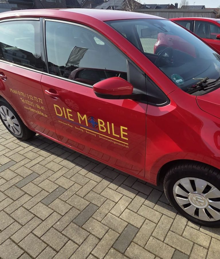 Die Mobile Zwei Frauen in roten Arztkitteln stehen neben einem weißen Auto mit Logo.